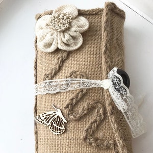 May include: A burlap clutch with a white crocheted flower, a wooden butterfly, and a lace ribbon. The clutch has a brown leather strap and a braided rope detail.