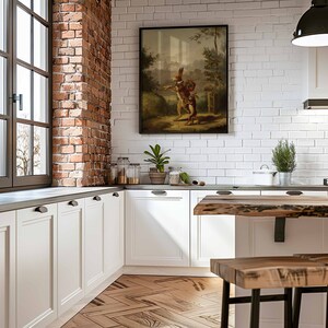 May include: A white kitchen with white cabinets and a brick accent wall. A framed painting of a rabbit with a bag over its shoulder and a smaller rabbit following behind is hanging on the wall. A wooden table with a live edge sits in the foreground.