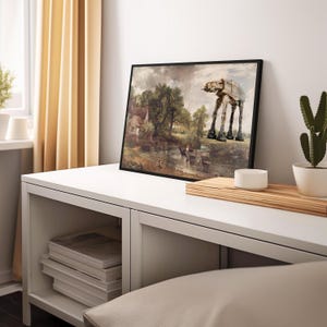 May include: Framed art print featuring a landscape painting with an AT-AT walker superimposed. The artwork has a black frame and depicts a rural scene with a river, trees, and a building. The AT-AT walker is a light tan color.