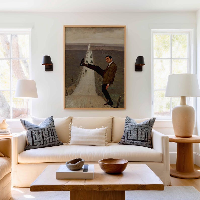 May include: A framed wall art print featuring a man in a brown blazer and dark trousers standing on a road, one leg raised high. The artwork is displayed above a cream-colored sofa with two patterned gray pillows and a central white lumbar pillow.