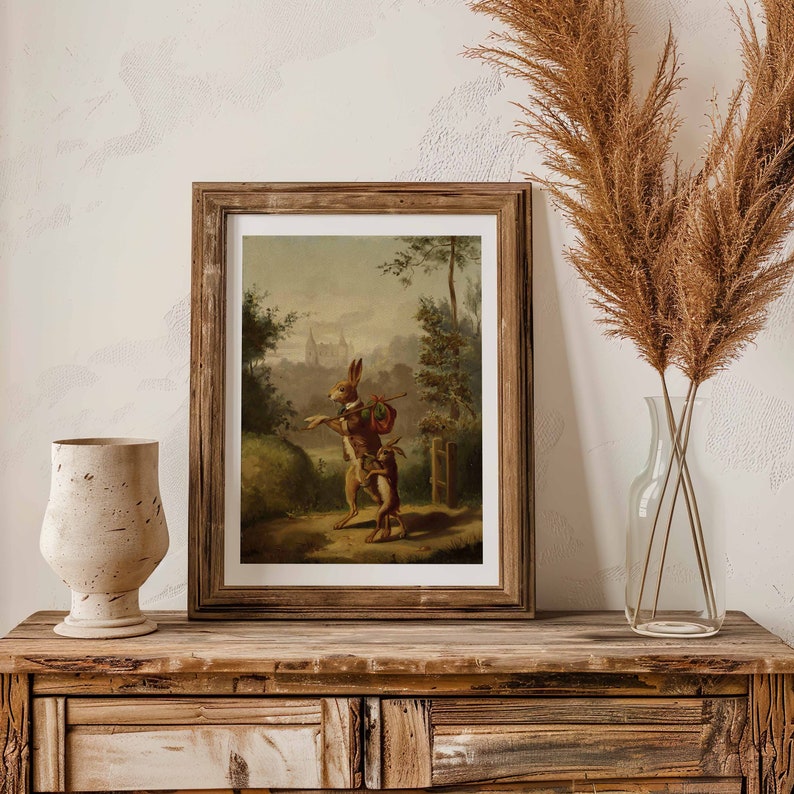 May include: A framed vintage illustration of two rabbits walking through a forest. The rabbit in the front is wearing a red coat and carrying a bag. The background is a forest scene with a distant town in the background.