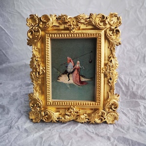 Op de afbeelding: Een gouden lijst met een schilderij van twee figuren die op een vis rijden. De figuren dragen rode en witte kleding. De achtergrond is blauwgroen.