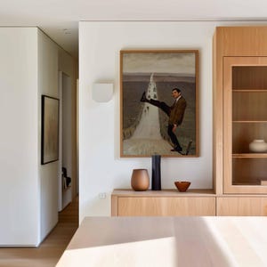 May include: A framed rectangular wall art piece depicting a man in a brown blazer and black trousers standing on a long, narrow road that stretches into a gray landscape. The artwork is displayed above a light wood cabinet with a vase and bowl.