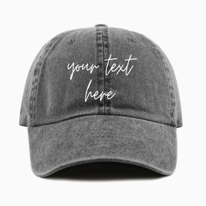 A charcoal baseball cap is shown with embroidered text saying 'your text here' on two lines in all lowercase and a cursive font with white thread. The baseball cap is pigment-dyed and features a faded/worn in look due to this.