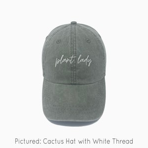 A cactus green baseball cap is shown with embroidered text saying "plant lady" in all lowercase and a cursive font with white thread. The baseball cap is pigment-dyed and features a faded/worn in look due to this.