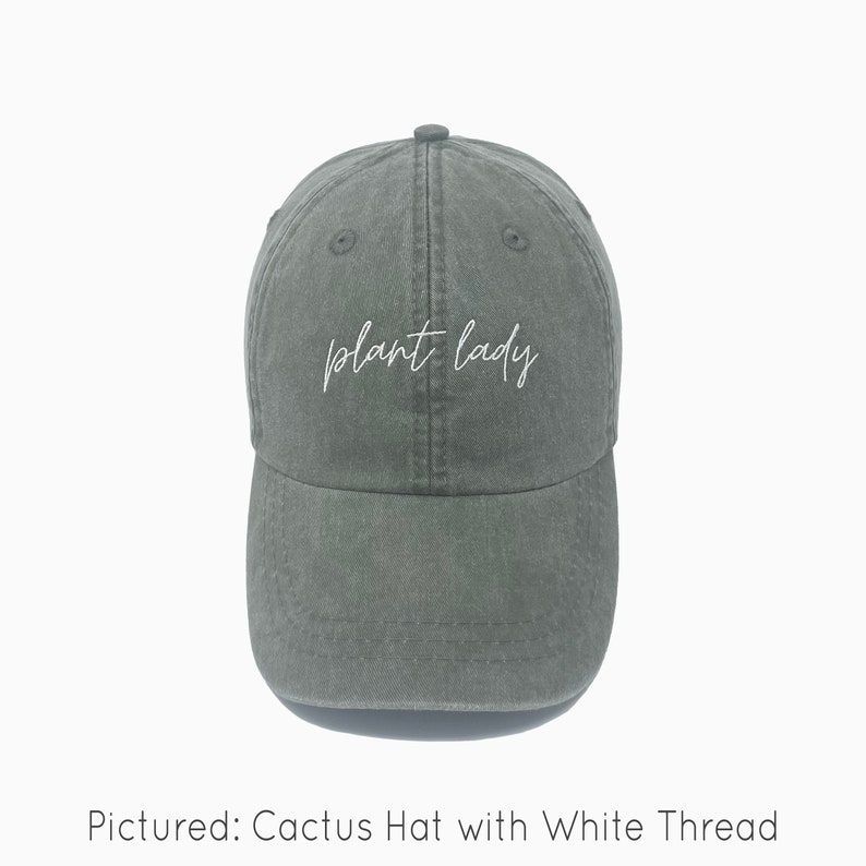 A cactus green baseball cap is shown with embroidered text saying "plant lady" in all lowercase and a cursive font with white thread. The baseball cap is pigment-dyed and features a faded/worn in look due to this.