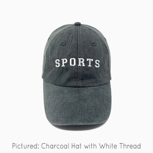 A charcoal gray baseball cap is shown with embroidered text saying "sports" in all uppercase in an athletic type font with white thread. The baseball cap is pigment-dyed and features a faded/worn in look due to this.