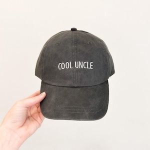May include: A charcoal gray baseball cap with a washed, vintage-style finish. The front of the cap features the text "COOL UNCLE" embroidered in white, sans-serif capital letters. The hat has a structured crown, a curved brim, and eyelets.