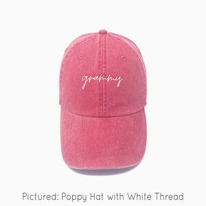 A poppy red baseball cap is shown with embroidered text saying "grammy" in all lowercase and a cursive font with white thread. The baseball cap is pigment-dyed and features a faded/worn in look due to this.