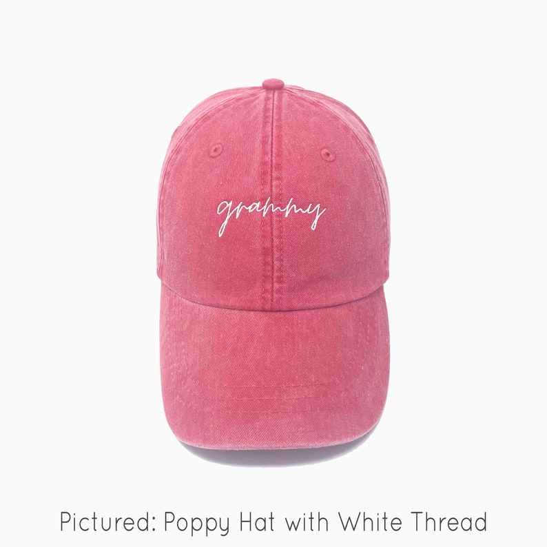 A poppy red baseball cap is shown with embroidered text saying "grammy" in all lowercase and a cursive font with white thread. The baseball cap is pigment-dyed and features a faded/worn in look due to this.