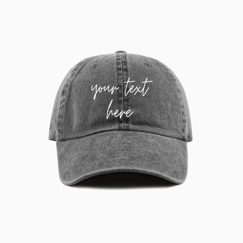 A charcoal baseball cap is shown with embroidered text saying 'your text here' on two lines in all lowercase and a cursive font with white thread. The baseball cap is pigment-dyed and features a faded/worn in look due to this.