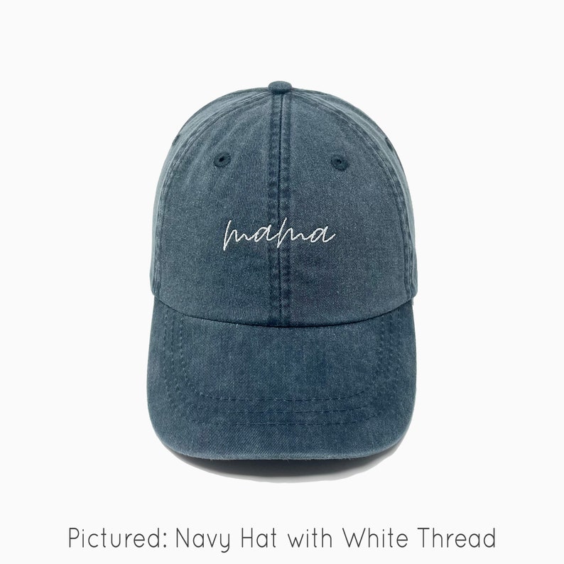 A navy baseball cap is shown with embroidered text saying "mama" in all lowercase and a cursive font with white thread. The baseball cap is pigment-dyed and features a faded/worn in look due to this.