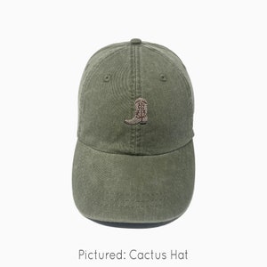 A cactus green baseball cap is shown with an embroidered cowboy boot. The boot is light tan with dark brown accents and a dark gray sole. The baseball cap is pigment-dyed and features a faded/worn in look due to this.