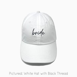 A white baseball cap is shown with embroidered text saying "bride" in all lowercase and a cursive font with black thread. The baseball cap is pigment-dyed and features a faded/worn in look due to this.