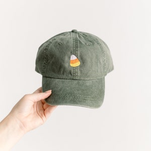 May include: A green baseball cap with a curved brim. The cap features an embroidered candy corn design in white, yellow, and orange. The hat is made of a soft, worn material.