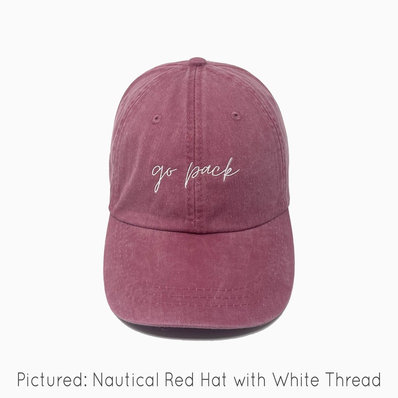 A nautical red baseball cap is shown with embroidered text saying "go pack" in all lowercase and a cursive font with white thread. The baseball cap is pigment-dyed and features a faded/worn in look due to this.