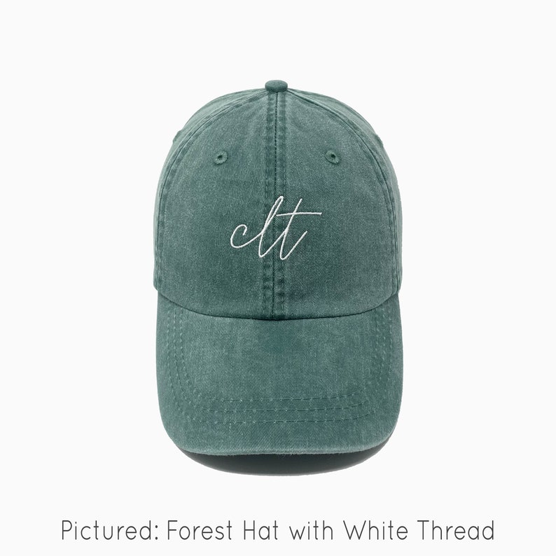 A forest green baseball cap is shown with embroidered text saying "clt" in all lowercase and a cursive font with white thread. The baseball cap is pigment-dyed and features a faded/worn in look due to this.