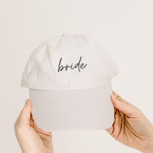 May include: A light gray baseball cap featuring the word bride embroidered in black cursive script on the front panel. The cap has a structured crown, a curved brim, and stitched eyelets for ventilation, providing a clean and simple aesthetic.
