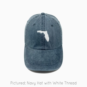 A navy baseball cap is shown with an embroidered image of the state of Florida in white thread. The baseball cap is pigment-dyed and features a faded/worn in look due to this.