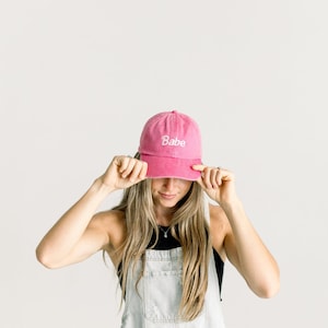 May include: A bright pink baseball cap featuring the word Babe embroidered in white script lettering on the front panel. The cap has a curved brim and a structured crown, constructed from a solid-colored fabric with matching stitching details.
