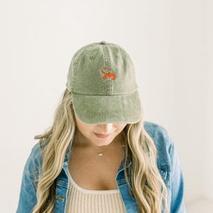 May include: A sage green baseball cap with a distressed, washed-out texture. The front of the cap features an embroidered design in bright orange thread, consisting of a stylized, cursive letter S with a small, curved accent mark above it.