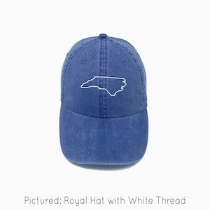 A royal blue baseball cap is shown with embroidered outline of the state of North Carolina in white thread. The baseball cap is pigment-dyed and features a faded/worn in look due to this.