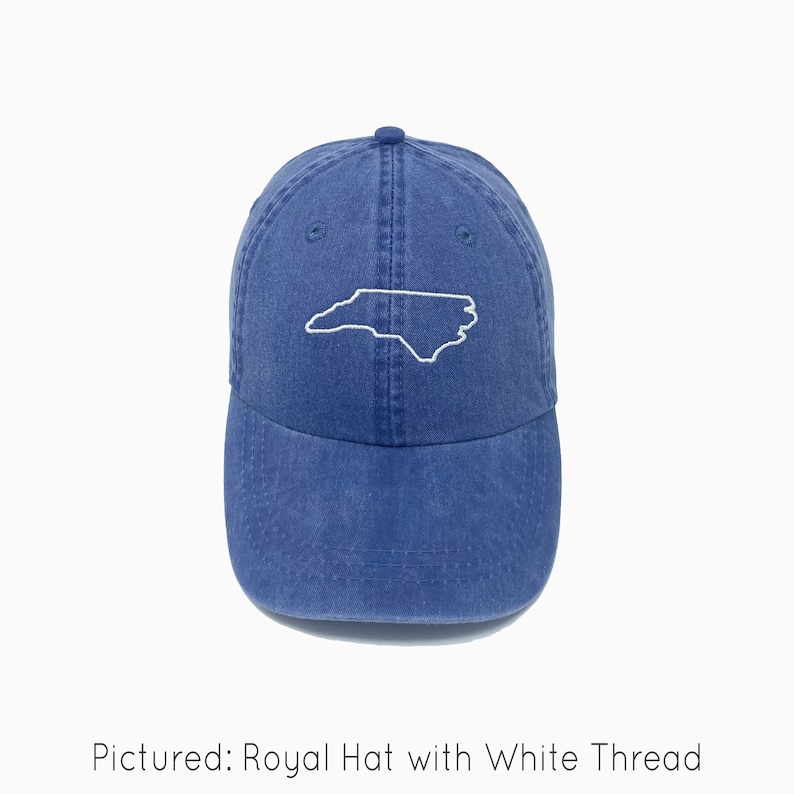 A royal blue baseball cap is shown with embroidered outline of the state of North Carolina in white thread. The baseball cap is pigment-dyed and features a faded/worn in look due to this.