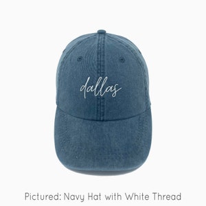 May include: A navy blue baseball cap with white embroidery that says "dallas" in a cursive font.