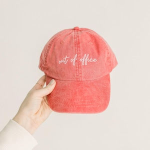 May include: A coral-colored baseball cap with a distressed, washed-out texture. The front panel features the text "out of office" embroidered in white cursive script. The cap has a curved brim and a button at the top center of the crown.