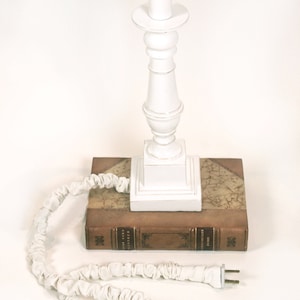 May include: White table lamp with a white fabric shade, sitting on top of a brown leather-bound book. The lamp has a white cord with a white plug.