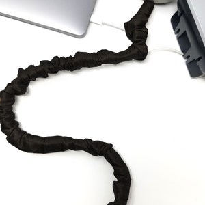 May include: A brown cord cover wraps around a white cable connecting a laptop to a power adapter. The cord cover is made of a soft, stretchy fabric and is designed to protect and organize cords. The text "COVER Ease decorative cord covers" is visible in the bottom right corner of the image.