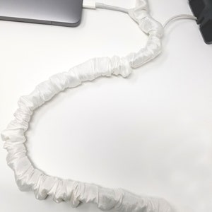 May include: White satin cord organizer for keeping cords and cables tidy. The cord organizer is wrapped around a white cable. The text "COVER Ease decorative cord covers" is visible at the bottom of the image.
