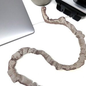 May include: A brown fabric cord organizer, shaped like a snake, is coiled on a white surface. The cord organizer is designed to keep cords and cables tidy and organized.