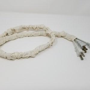May include: A white fabric cord organizer with three white electrical plugs attached.