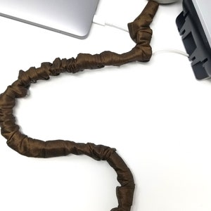 Może przedstawiać: Brązowy pokrowiec na kabel z tkaniny owinięty wokół białego kabla. Kabel jest podłączony do laptopa i zasilacza. Tekst "COVER Ease decorative cord covers" jest widoczny w prawym dolnym rogu obrazu.