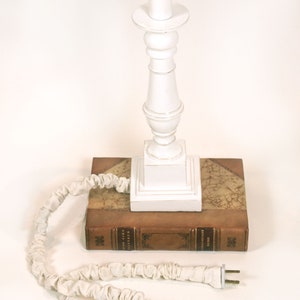 May include: White table lamp with a white fabric shade and a white base. The lamp is sitting on top of a brown leather-bound book. The lamp cord is white and has a white fabric cover.