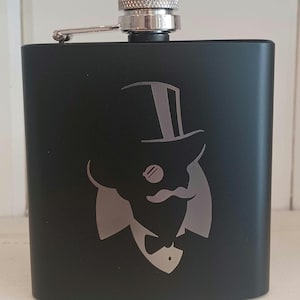 May include: Black metal flask with a silver cap and a silver engraved image of a man wearing a top hat, monocle, and bow tie.