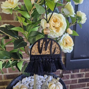 May include: A black macrame car charm with a leopard print design and the letter "M". The charm is hanging from a black cord with wooden beads. The charm is displayed in front of a plant with yellow flowers and small pumpkins.