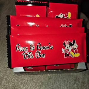 Puede incluir: Una caja de bolsas de regalo rojas y negras con los personajes de Mickey Mouse y Minnie Mouse. Las bolsas están decoradas con lunares blancos y el texto "Alex & Anna's Big One".