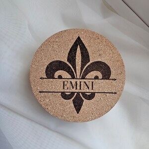 Puede incluir: Un posavasos de corcho con un diseño de flor de lis negro y el nombre "EMINI" grabado en el centro.