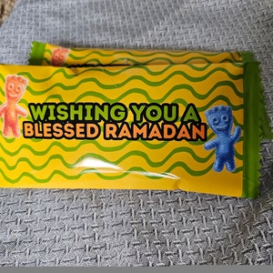 May include: A yellow and green candy wrapper with a wavy pattern. It features two cartoon gummy bears, one orange and one blue. The wrapper says "Wishing you a Blessed Ramadan".