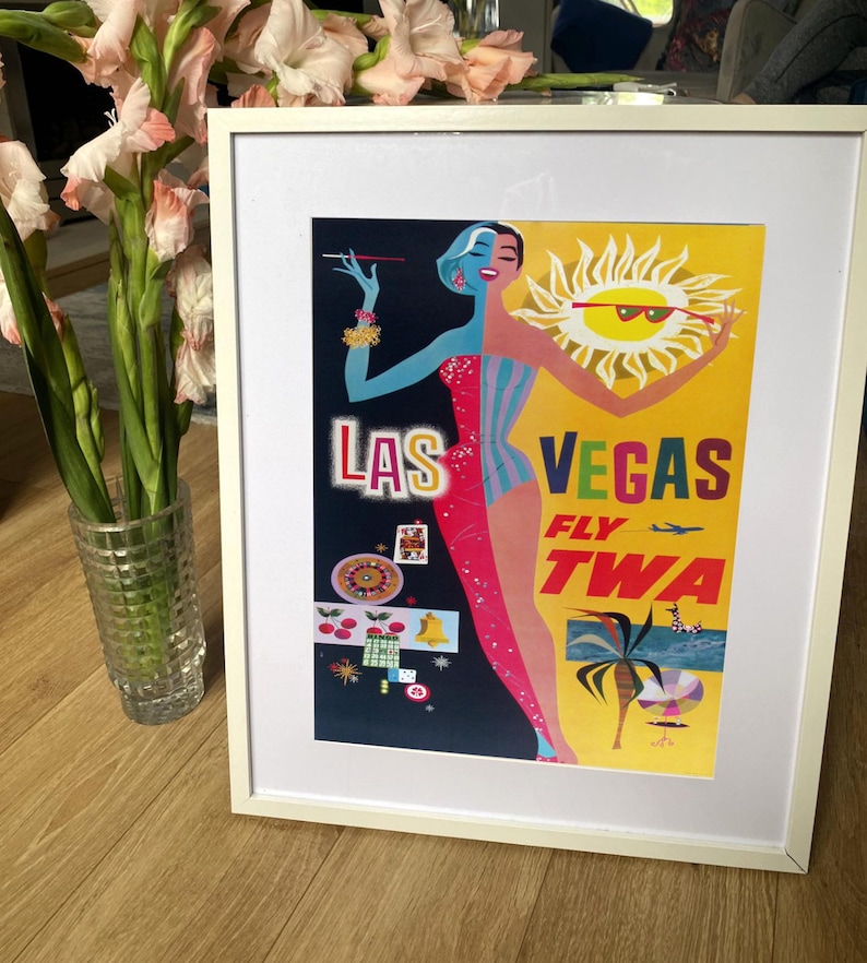 Op de afbeelding: Een vintage reisposter die reclame maakt voor een reis naar Las Vegas. De poster toont een vrouw in een rood-blauwe jurk, een gele zon met een zonnebril en de tekst "Las Vegas Fly TWA".