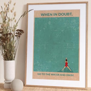 May include: A framed art print with the text "WHEN IN DOUBT, GO TO THE WATER AND SWIM." The print shows a swimmer in a red swimsuit against a teal background. The artwork is in a wooden frame and is displayed with a vase of dried flowers.
