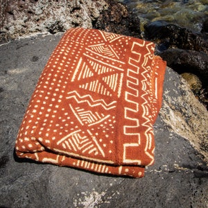 Puede incluir: Textil plegado de color óxido con un patrón geométrico crema. El diseño incluye puntos, zigzags y formas angulares. El textil está sobre una roca gris oscura cerca del agua.