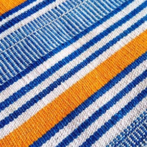 May include: Close-up of a woven textile with a striped pattern. The design features alternating bands of blue, white, and orange. The fabric has a textured appearance, with visible threads and a diagonal weave.