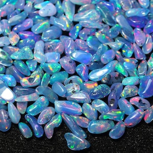 May include: A close-up of a collection of opals. The opals are in various shapes and sizes, with a range of colours including blues, greens, and purples. The gemstones have iridescent flashes and are scattered on a dark surface.
