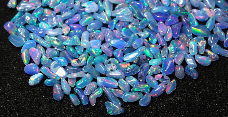 May include: A close-up of a collection of opals. The opals are in various shapes and sizes, with a range of colours including blues, greens, and purples. The gemstones have iridescent flashes and are scattered on a dark surface.