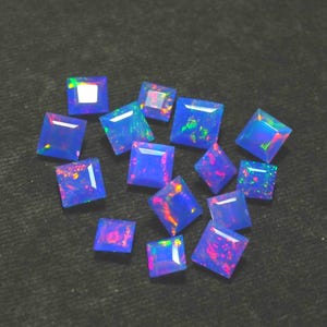 Op de afbeelding: Een verzameling vierkant geslepen, levendige blauwe opalen met flitsen van rood, groen en geel. De edelstenen liggen verspreid op een donkere ondergrond, waarbij hun gepolijste facetten en interne kleurenspel worden getoond. Geschikt voor sieraden.