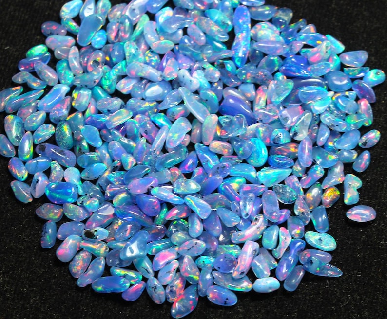 May include: A collection of polished opal gemstones in various shapes and sizes. The opals display a range of colours, including blues, greens, and flashes of pink and orange. The gemstones are scattered across a dark surface.