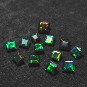 Puede incluir: Una colección de ópalos negros de corte cuadrado con vibrantes destellos verdes, azules y naranjas. Las piedras preciosas están dispersas sobre una superficie gris oscuro, mostrando sus cualidades iridiscentes. Adecuado para la creación de joyas.